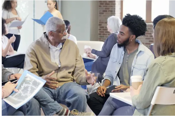 An older man and a younger man sit among a discussion circle; the older man holds a paper.